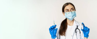 Covid-19 vaccination and healthcare concept. Young doctor in medical mask showing syringe and vaccine from coronavirus omicron variant, standing over white background.