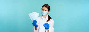 Portrait of medical worker, woman doctor in suggesting face masks from covid-19 pandemic, standing in hospital uniform over torquoise background.