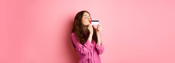 Image of young beautiful woman kissing her credit card, going on shopping, wasting money in stores, standing against pink background. Copy space