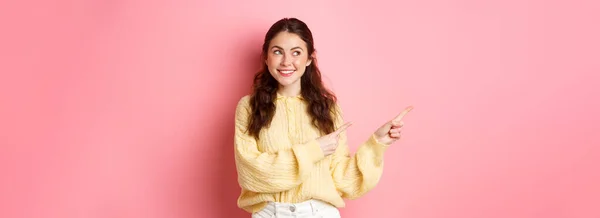 年轻快乐的女人微笑着 指指着左边的版面 望着左边的版面 展示着广告 站在粉色的春光背景下 — 图库照片