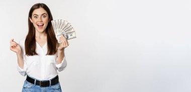 Enthusiastic woman showing money, cash and smiling, got loan, micro credit, standing delighted against white background.