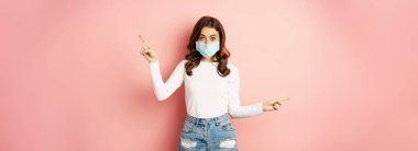 Portrait of beautiful woman in face medical mask pointing sideways, showing two choices, ways, sides, demonstrating advertisement, pink background.