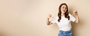 Self-assured young happy woman pointing fingers at herself and dancing, self-promoting, being confident, standing over beige background.