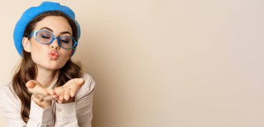 Beautiful stylish woman in sunglasses, blowing air kiss on hands near lips, gazing softly and flirty at camera, posing against beige background.