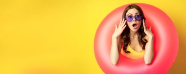 Portrait of young pretty woman in sunglasses, wearing beach swimming ring, looking surprised and amazed, shocked wow face, yellow background.