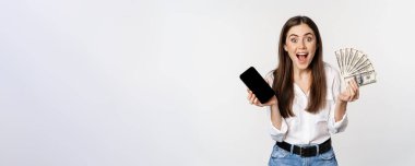 Enthusiastic young woman winning money, showing smartphone app interface and cash, microcredit, prize concept, standing over white background.