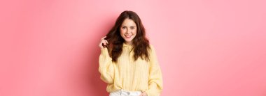 Güzel kız saçlarıyla oynuyor, gülümsüyor ve kameraya kendinden emin bir şekilde bakıyor, cazibe pozu veriyor, pembe arka planda duruyor..