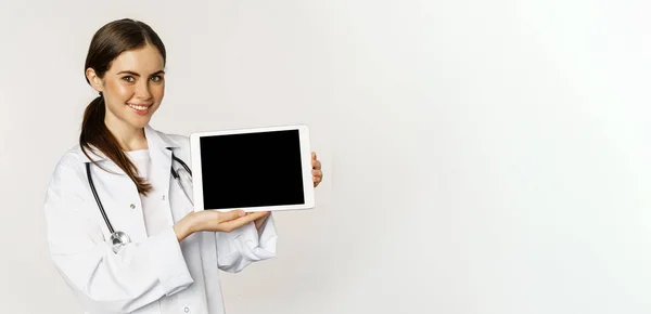 stock image Image of woman doctor, female healthcare worker showing online medical website, digital tablet screen and smiling, standing in white coat over white background.