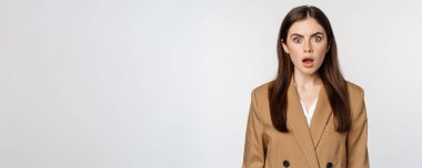 Portrait of shocked, speechless businesswoman gasping, staring stunned at camera, wearing brown suit, white background.