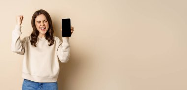Happy woman winning, showing smartphone screen and shouting of joy, fist pump, standing over beige background.
