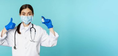 Gülümseyen doktor portresi, tıbbi personel yüz maskesi ve lastik eldiven, baş parmak ve koronavirüs gösteren, omikron aşısı, mavi arka planda duran.