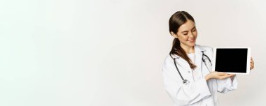 Smiling doctor, female physician or nurse showing digital tablet screen, demonstrating online website, application, standing over white background.