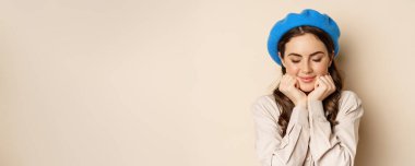 Close up portrait of feminine young woman in trendy french hat, posing cute and romantic, making silly coquettish face and tender smile, beige background.