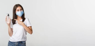 Covid-19, sağlık ve sosyal uzaklık kavramı. Tıbbi maske takan sevimli esmer kadın el dezenfektanı kullanıp şişeyi işaret ediyor..
