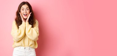 Heyecanlı güzel kız şaşırmış ve şaşkın şaşkın bakıyor, yanaklarından elele tutuşuyor ve nefes nefese, promosyon teklifine şaşırmış bir şekilde bakıyor, fotokopi alanı pembe arka planının yanında duruyor..