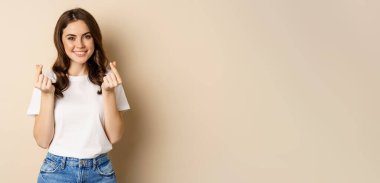 Cute young woman showing finger hearts sign and smiling, express care and love, standing over beige background.