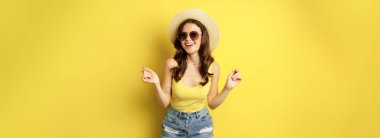 Stylish girl in straw hat and tank top, ready for summer, going on vacation and smiling pleased, standing over yellow background.