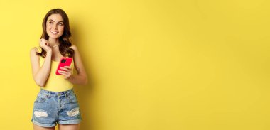 Technology and cellphone. Beautiful stylish girl order in online store, praying in application, using smartphone app and thinking, making decision, standing over yellow background.
