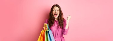 Heyecanlı kız sevinç çığlığı, yumruk pompası yapıyor, alışveriş torbası tutuyor ve seviniyor, pembe arka planda elbise içinde duruyor..