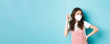 Covid, sağlık ve salgın hastalık konsepti. - Çok iyi. SağIık maskesi takan genç destekçi bir kadın onaylama işareti gösteriyor. Herkesin içinde maske takıyor. Mavi arka plan..