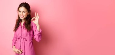 Tatmin olmuş genç bir kadın doymuş hissediyor, iyi yedikten sonra koca göbeğine dokunuyor, onay işareti gösteriyor, harika bir yemek, pembe arka plan..