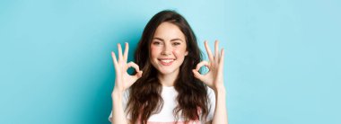 Kendine güvenen gülümseyen bir kadının portresi tamam diyor, tamam diyor, size katılıyor, onaylıyor ve tavsiye ediyor, mavi arka planda duruyor..