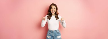 Enthusiastic beautiful and stylish girl showing advertisement, pointing fingers down at announcement and smiling, pink background.