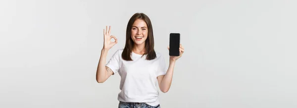 笑顔でくださいブルネットの女の子はスマートフォンの画面を表示し 承認で良いジェスチャーを示します — ストック写真