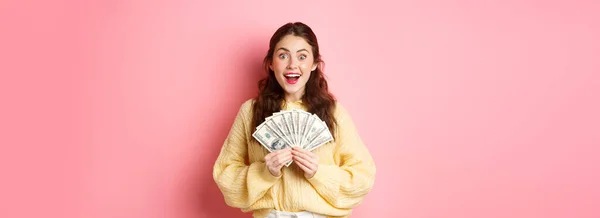 Wow I won. Excited brunette girl showing money and smiling, got her cash prize, holding dollar bills and looking happy, standing over pink background.