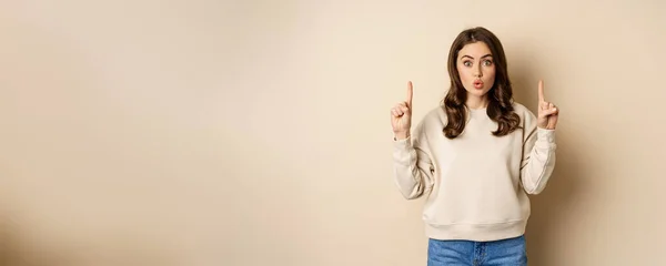 stock image Surprised beautiful girl showing advertisement, pointing fingers on top, store sale or website link up, standing over beige background.