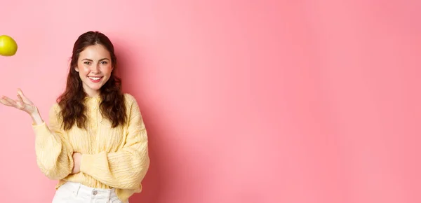 stock image Young smiling woman looking with confidence, throwing apple in air, eating healthy fruits to keep that smile perfect, standing against pink background.