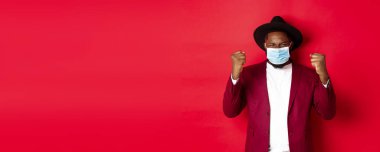 Covid-19, karantina ve tatil konsepti. Neşeli Afrikalı Amerikalı adam yumruğunu sıktı ve kazandığı için sevindi, amacına ulaştı, Coronavirus 'un tıbbi maskesini taktı..