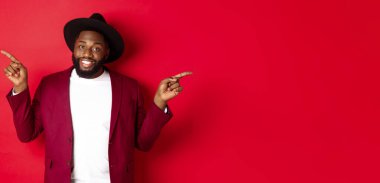 Kış tatili ve alışveriş konsepti. Neşeli siyahi adam gülümsüyor ve iki reklam gösteriyor, fotokopi alanlarını işaret ediyor, kırmızı arka plan.