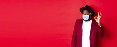 Covid 19 ve moda konsepti. Şapkalı ve ceketli şık Afro-Amerikan adam, yüz maskesi takıyor ve kırmızı arka planda duruyor..