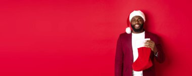Yakışıklı Afro-Amerikan adam kış tatilini kutluyor, Noel çorabı giyiyor ve gülümsüyor, Noel Baba şapkası takıyor, kırmızı arka planda duruyor..
