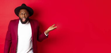 Kızgın ve kafası karışık siyah adam tartışıyor, kameraya bağırıyor ve elini kaldırıyor. Bir şey anlamıyorum, kırmızı arka plan..