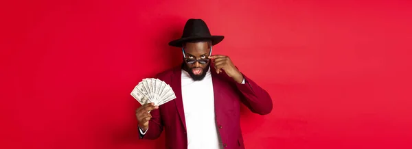 Sassy and cool Black man in hat and party outfit, showing dollars and looking from under sunglasses, standing over red background.