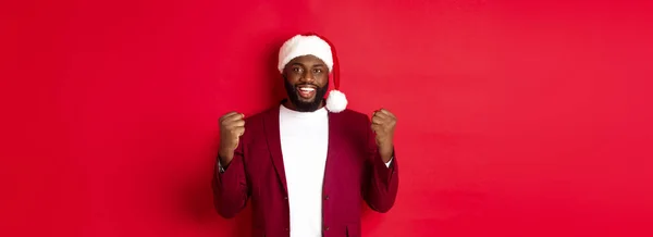 stock image Christmas, party and holidays concept. Excited Black man celebrating New Year, wearing santa hat, clench fists satisfied, winning and rejoicing, standing against red background.