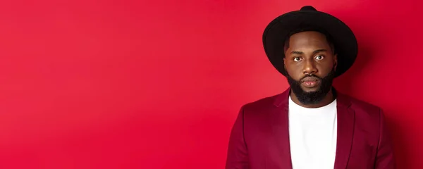 stock image Close-up of serious-looking african american guy in blazer and black hat, looking at camera, standing over red background.