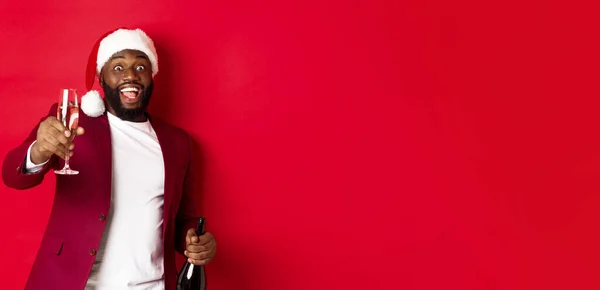 Christmas, party and holidays concept. Handsome Black man in santa hat raising glass of champagne and smiling, saying toast, celebrating New Year, red background.