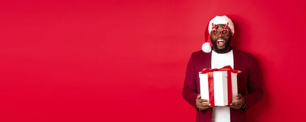 stock image Christmas. Handsome african american man in party glasses and santa hat holding new year gift, looking surprised, receive present and smiling, standing over red background.