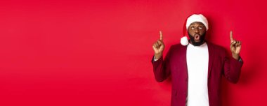 Noel, parti ve tatil konsepti. Etkilenmiş siyah adam Noel Baba şapkalı, işaret ediyor, hayret ediyor ve kameraya bakıyor, tanıtım teklifini kontrol ediyor, kırmızı arka plan.