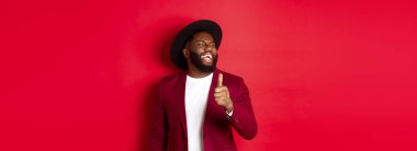 Yakışıklı Afro-Amerikan erkeği eğleniyor, başparmağını kaldırıyor ve iyi bir şakadan gülüyor, kırmızı arka planda duruyor..