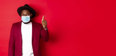 Covid 19 ve moda konsepti. Şapkalı ve ceketli şık Afro-Amerikan adam, yüz maskesi takıyor ve başparmağını kaldırıyor, kırmızı arka planda duruyor..