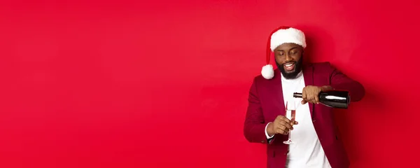 Christmas, party and holidays concept. Handsome Black man in santa hat pouring glass of champagne and smiling, celebrating New Year, red background.