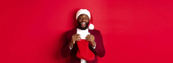 stock image Excited black man open christmas sock with presents and sweets, smiling happy, standing in santa hat against red background.