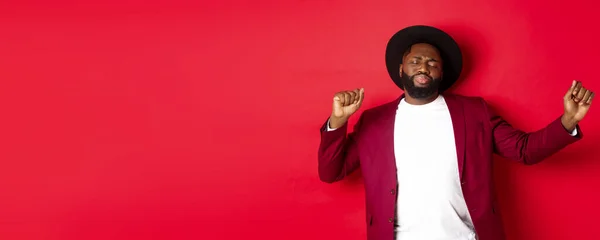 stock image Christmas and people concept. Happy Black man celebrating New Year and dancing, wearing party outfit, red background.