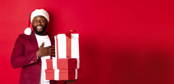 Christmas, New Year and shopping concept. Happy Black man receiving xmas gifts, saying thank you and smiling grateful, standing in santa hat against red background.