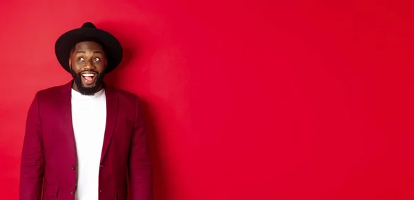 stock image Fashion and party concept. Excited african american man looking at logo, staring upper left corner with happy smile, standing against red background.