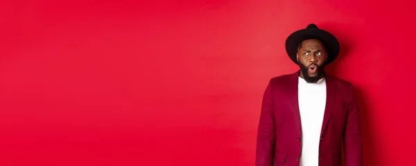 Fashion and party concept. Shocked and displeased black man staring at something unpleasant, looking upper left corner and frowning, standing against red background.